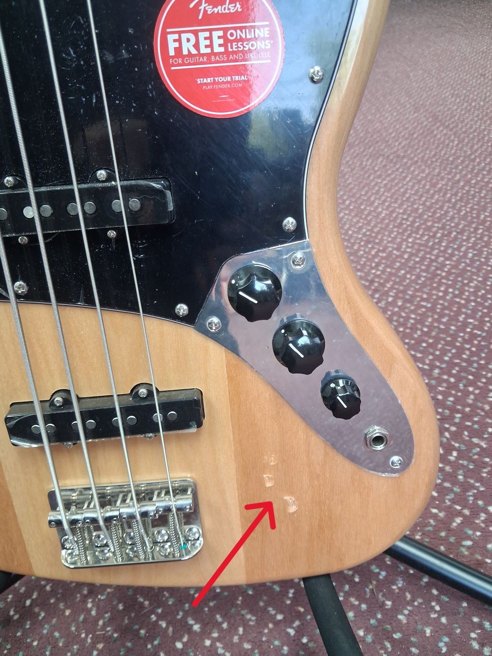 Bass guitar with a Fender sticker on a patterned surface with an arrow pointing to some small indentations on the lower body