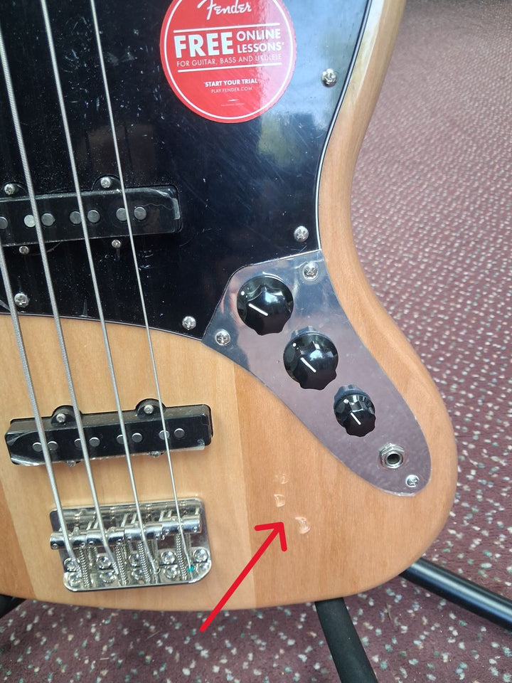 Bass guitar with a Fender sticker on a patterned surface with an arrow pointing to some small indentations on the lower body