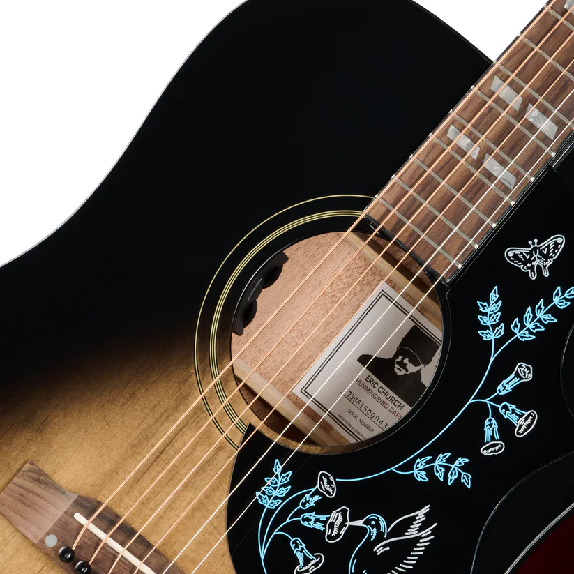 Close-up of an acoustic guitar body and neck showing a black-to-brown sunburst finish, light wood soundboard, blue floral inlay on a black pickguard.