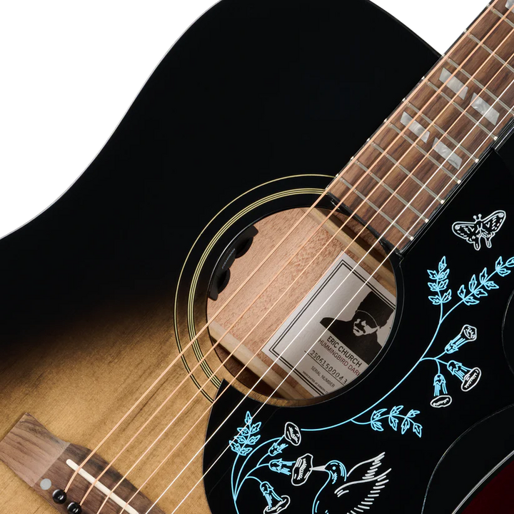 Close-up of an acoustic guitar body and neck showing a black-to-brown sunburst finish, light wood soundboard, blue floral inlay on a black pickguard.
