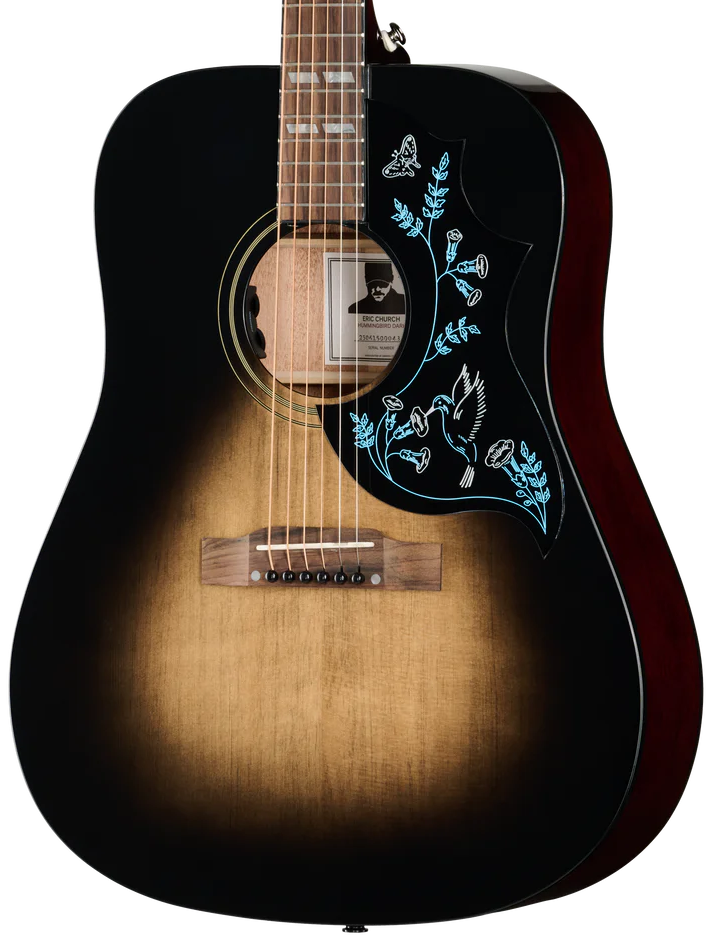An acoustic guitar with a black-to-brown sunburst finish, featuring light blue floral and bird inlay on the pickguard