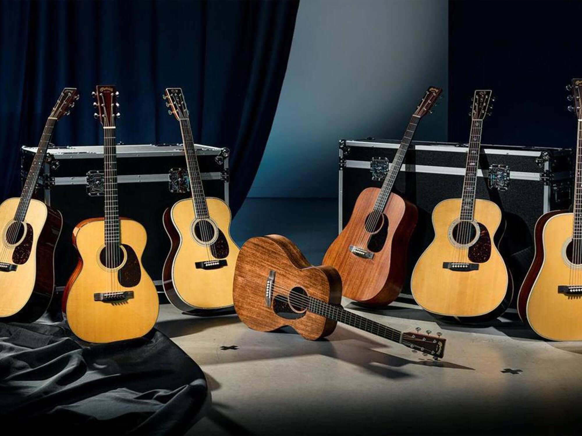 A lineup of Martin acoustic guitars in various natural wood finishes, with some standing upright on stands and one lying diagonally in the foreground, set against a dark blue curtain backdrop.