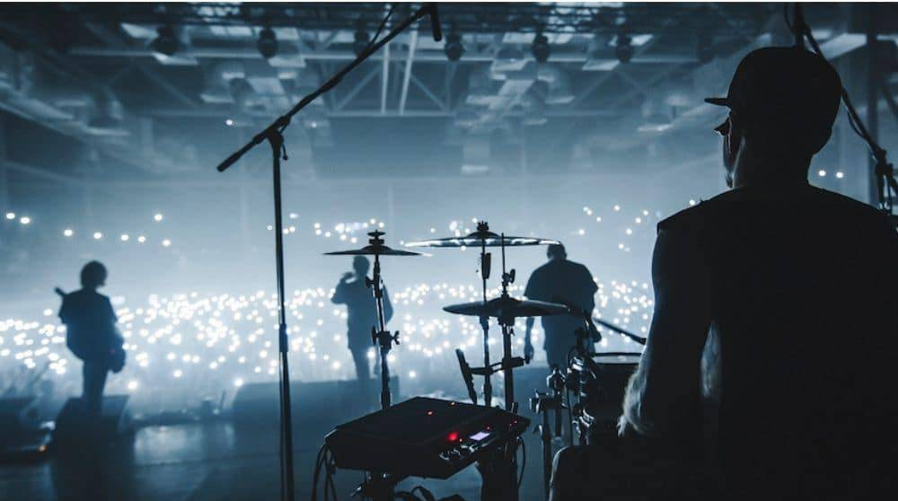 A dark concert stage scene with band members in silhouette, including a drummer and keyboardist, with a large crowd of people holding up lights in the background.